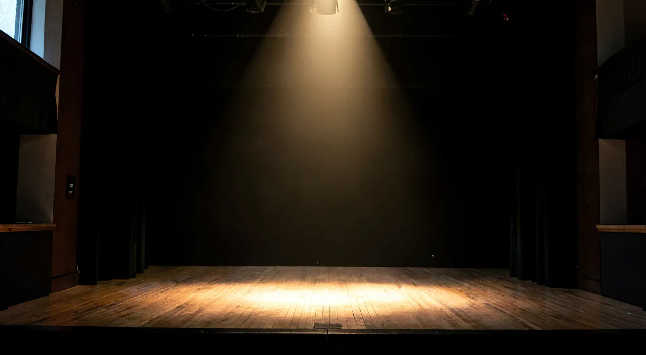 An empty theatre stage lit by a single warm spotlight, dark wings either side, before the November season begins